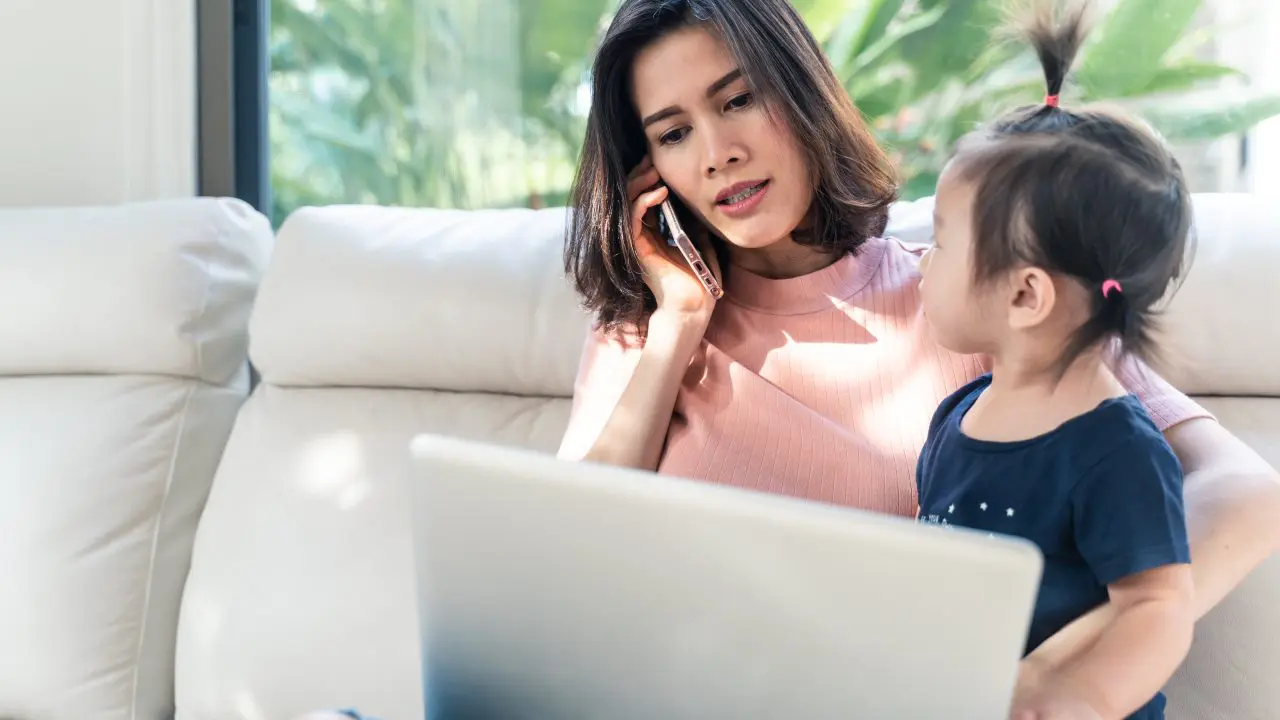 Asian mum on the phone with young child and laptop in front of her.