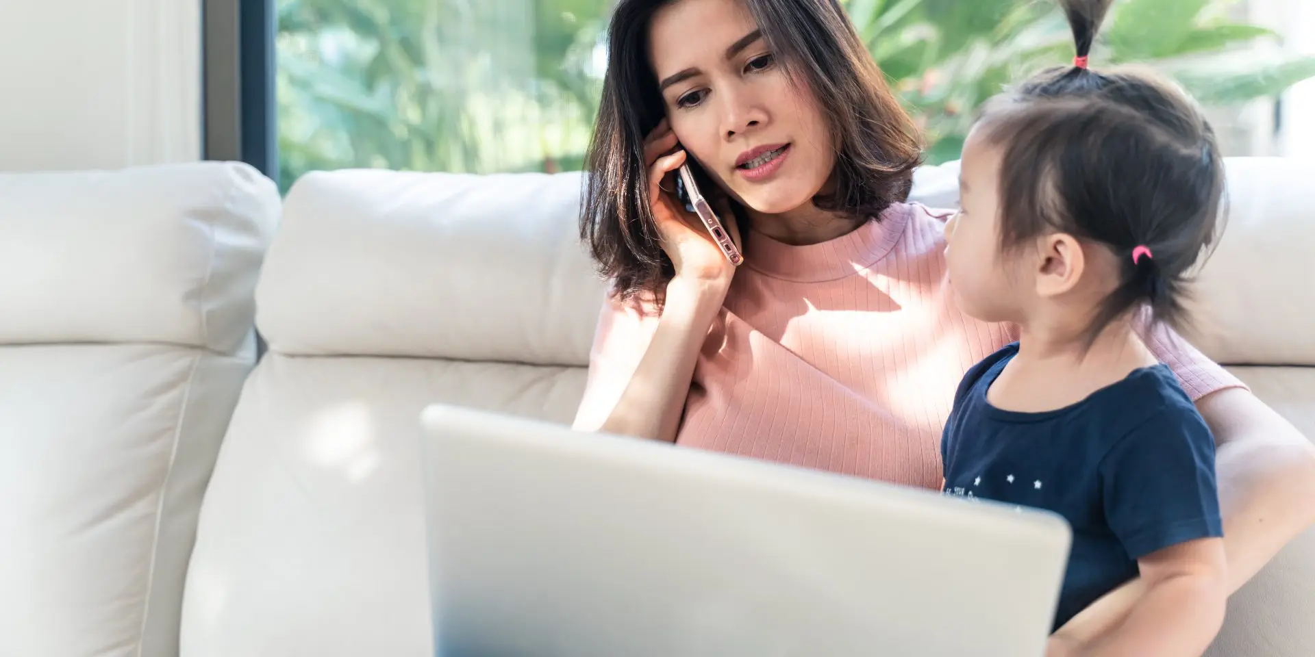 Asian mum on the phone with young child and laptop in front of her.