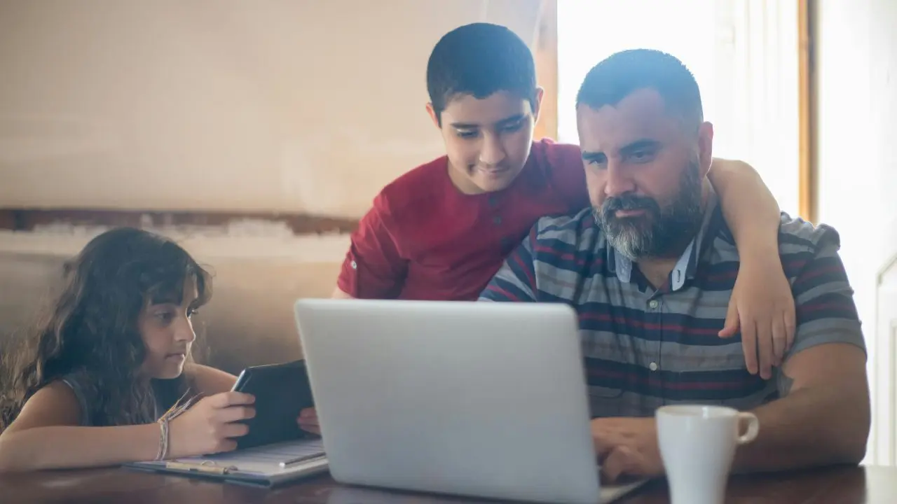 Dad using laptop with son watching on and daughter using tablet device.
