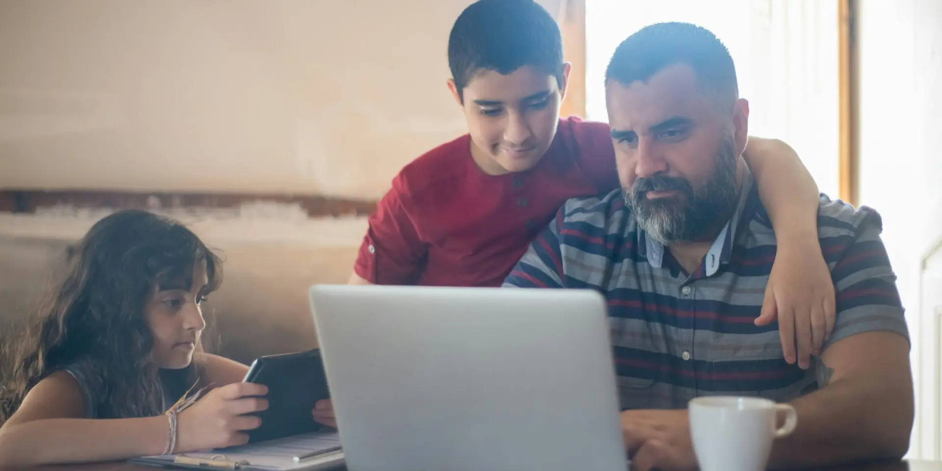 Dad using laptop with son watching on and daughter using tablet device.