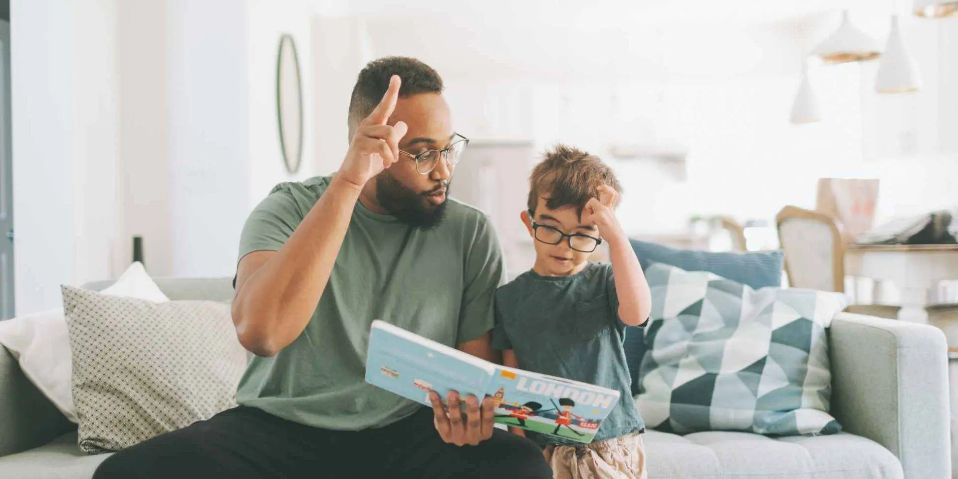Dad and son sat on a sofa reading a book together and making hand gestures