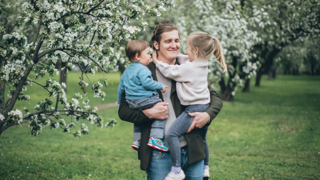 A smiling man stands by blossom trees holding his two young children in his arms.