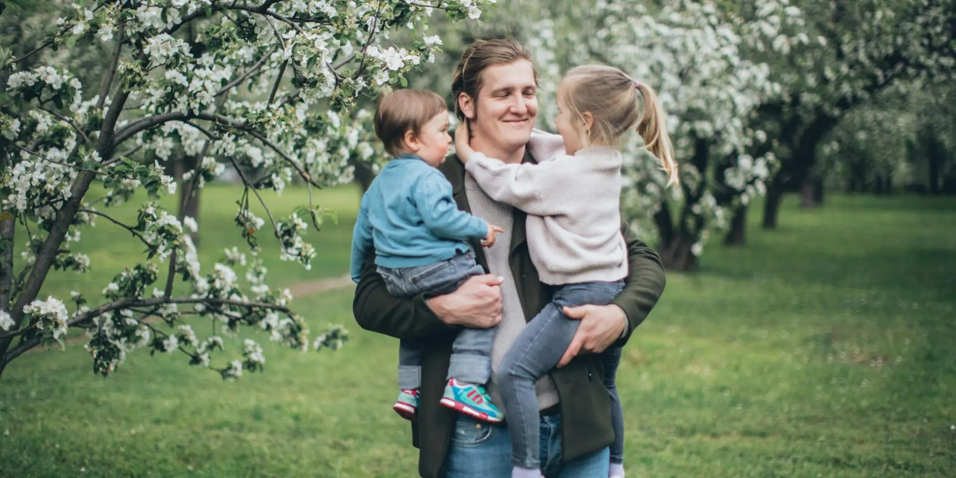 A smiling man stands by blossom trees holding his two young children in his arms.