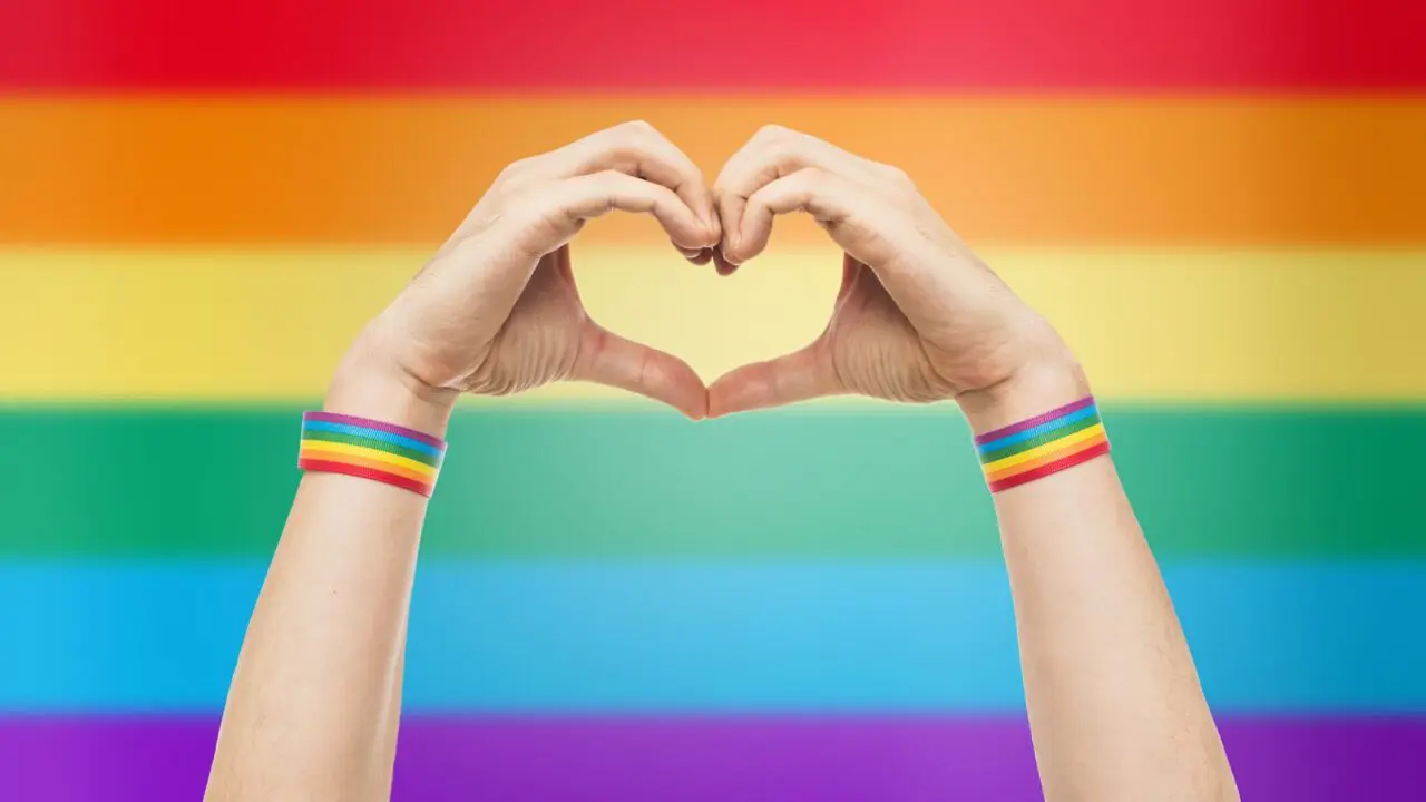 Hands creating a love heart with a rainbow flag in the background to represent support for the LGBTQIA+ community