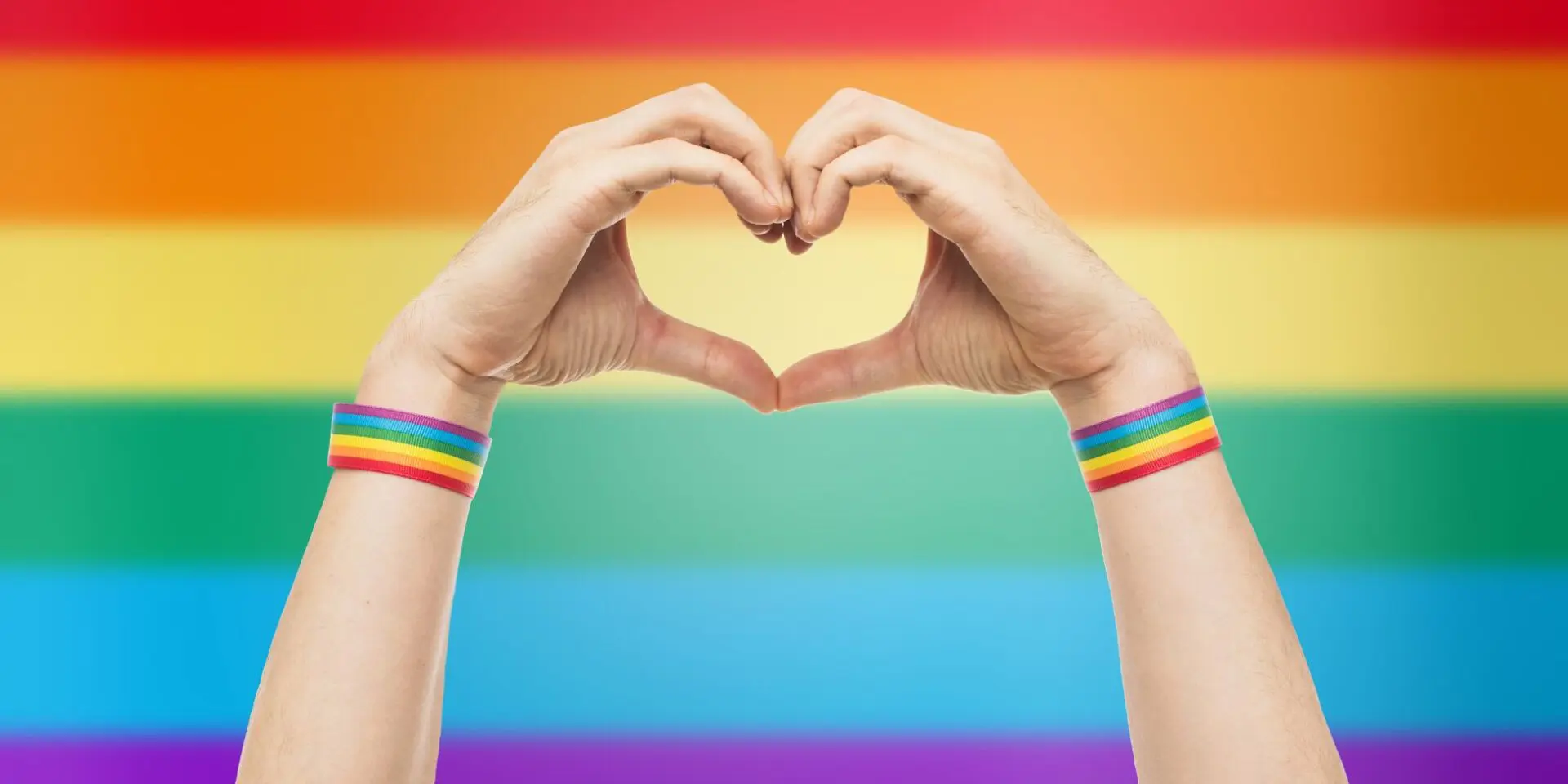 Hands creating a love heart with a rainbow flag in the background to represent support for the LGBTQIA+ community